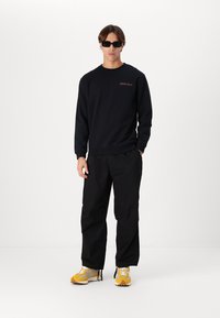 Zwarte sweatshirt met rood logo, gecombineerd met losse zwarte broek en geel/grijze sneakers. Model draagt zwarte zonnebril.