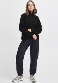Zwarte gebreide coltrui met geribbelde textuur, gecombineerd met marineblauwe cargobroek en lichte sneakers met grijze en crème accenten.
