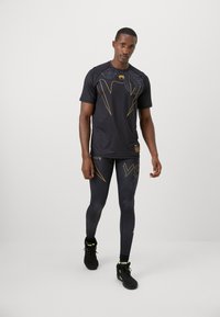 Hombre caminando vestido con un atuendo atlético negro con diseños geométricos dorados y de serpiente, y zapatillas negras con acentos en verde neón.