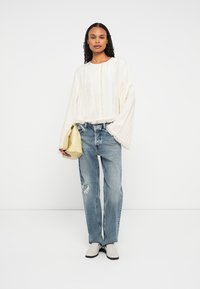Frau trägt ein lockeres, weißes, strukturiertes Langarmshirt, zerrissene blaue Jeans, weiße Schuhe und hält eine gefaltete gelbe Handtasche.
