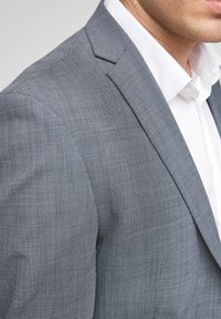 Grijze geruite blazer met een getailleerde pasvorm, gestructureerde schouders en rechtse revers, gecombineerd met een witte overhemden met kraag eronder.