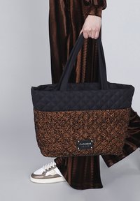 Persoon die een zwart en oranje textuur tote bag met "Cafenoir" label vasthoudt, gekleed in bruine zijden pants en witte sneakers.