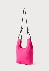 Jasnoróżowa torebka na ramię o gładkiej fakturze, wyposażona w czarne regulowane paski zawiązane w supełki. Prosty, funkcjonalny design.