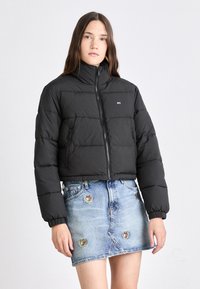 Svart puffjakke med høy krage, glidelåslukking og elastiske ermer. Båret over et blått denimskjørt med broderte lapper.