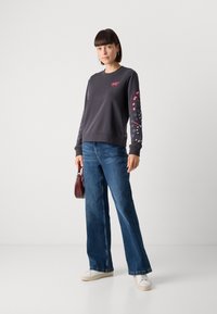 Felpa grigia con toppe a forma di cuore rosse, maniche a fantasia. Abbinata a pantaloni jeans a gamba larga blu e sneakers bianche, mentre tiene in mano una borsa bordeaux.