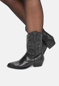 Botas de cowboy de cuero negro con puntas afiladas, detalles de costura en el eje y tacones bajos en bloque. Cremallera en la parte posterior para facilitar el uso.