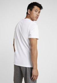 Hvid bomulds t-shirt med korte ærmer, rund halsudskæring og en afslappet pasform, parret med grå shorts. Modellen er vendt let mod kameraet.