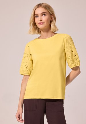 Lyshåret kvinde iført en gul bluse med broderede puffærmer og mørkebrune bukser, stående mod en ensfarvet baggrund.