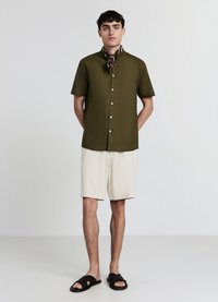 Olivgrünes Kurzarmhemd mit Stehkragen, kombiniert mit beigen Shorts und schwarzen Slip-On-Sandalen. Enthält einen braunen gemusterten Schal.