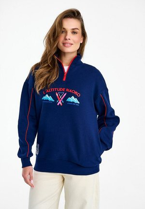 Kobieta ubrana w granatową bluzę z zamkiem do połowy długości i czerwonymi wykończeniami oraz grafiką z motywem narciarskim, stojąca na jednolitym białym tle.