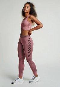 Set sportivo rosa chiaro composto da reggiseno e leggings con dettagli testurizzati e motivi a chevron neri; modella con scarpe sportive bianche.
