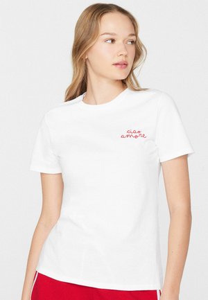 Junge Frau trägt ein weißes T-Shirt mit rotem "ciao amore" Schriftzug auf der Brust und rote Hose, schaut nach links vor einfachem Hintergrund.
