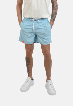 Uomo che indossa pantaloncini azzurri con coulisse, sneakers bianche e una camicia bianca con tatuaggi visibili su braccia e gambe.