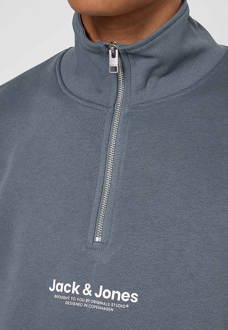 Grau halbzip Pullover aus strukturiertem Stoff, mit einem erhöhten Kragen, silbernem Reißverschluss und weißem, aufgedrucktem Logo auf der Vorderseite.