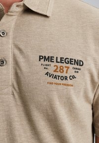 Beige polo tričko s texturovanou tkaninou. Má límec, tři knoflíky a černý a oranžový potištěný text značky na hrudi.