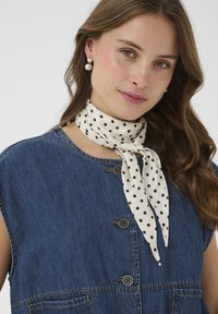 Femeie cu păr ondulat castaniu poartă cercei cu perle, o vestă din denim albastră fără mâneci și un fular alb cu buline legat în jurul gâtului.