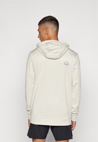 Krēmskrāsas kapucīte ar aukliņu, garām piedurknēm un mazu logotipu augšējā aizmugurē. Mīksta auduma, brīva piegriezuma, apvienota ar tumšiem šortiem.