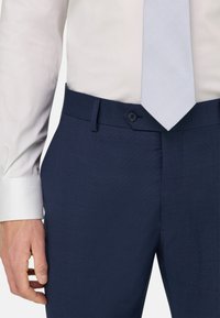 Homme portant un pantalon de costume bleu marine, une chemise blanche et une cravate bleu clair à motifs, la main reposant le long du corps.