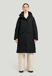 Svart ytterjacka med huva, med en placket-design, breda ärmar och framfickor. Bärs över en vit polotröja och ljusa byxor.