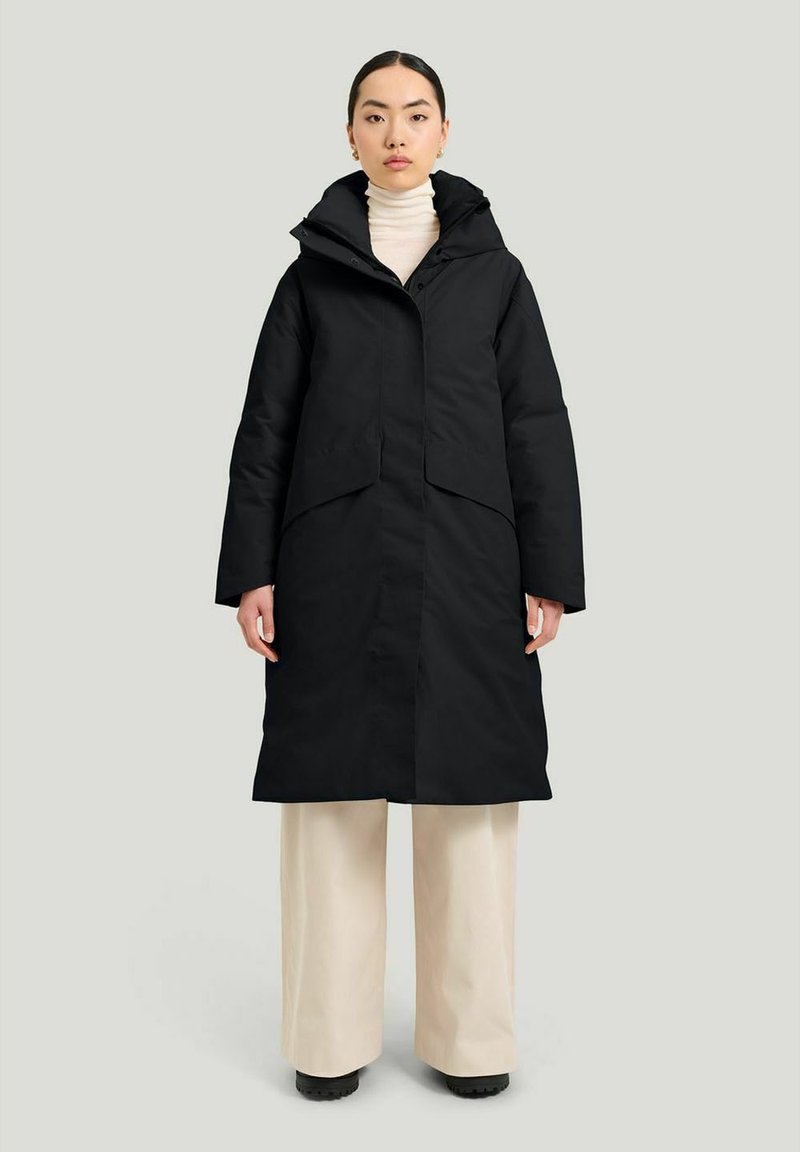 Svart ytterjacka med huva, med en placket-design, breda ärmar och framfickor. Bärs över en vit polotröja och ljusa byxor.