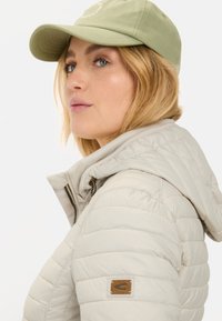 Licht beige pufferjack met een capuchon, met een subtiele quilted textuur en een bruin logo-label op de mouw. Gecombineerd met een lichtgroene pet.