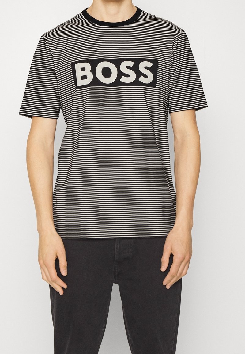Homme portant un t-shirt à rayures horizontales noires et blanches avec le mot « BOSS » en lettres blanches en gras sur un rectangle noir sur la poitrine.