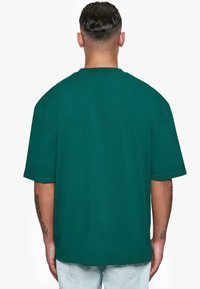 Camiseta de gran tamaño en color teal, de manga corta, con cuello redondo, hecha de algodón, presenta una textura suave y costuras visibles a lo largo del dobladillo.