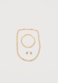 Σετ κοσμημάτων σε χρυσό τόνο με διαυγή στρογγυλά κρύσταλλα, που περιλαμβάνει κολιέ, βραχιόλι και αντιστοιχισμ ένα καρφωτά σκουλαρίκια τοποθετημένα σε λευκό φόντο.