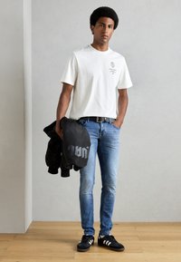T-shirt bianca in cotone con logo, giacca bomber nera con testo grigio, jeans azzurri chiari e sneakers Adidas nere con strisce bianche.