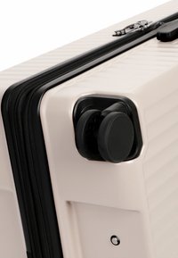 Primo piano dell'angolo di una valigia rigida beige che mostra le doppie ruote girevoli nere e la cerniera nera.