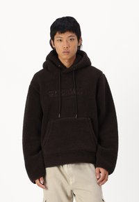 Hnědý fleecový hoodie s tkaničkami, měkkou texturou, přední kapsou a embosovaným logem "ORIGINALS". Volný střih a dlouhé rukávy.