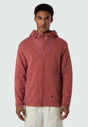 Hombre con una chaqueta con capucha color rosa polvo con cremallera y bolsillos delanteros, y pantalones de color claro, de pie contra un fondo sencillo.