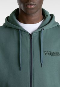 Grön huva med framzipper, snodd och broderad "vans"-logotyp. Slät tygtextur med en åtsittande design runt nacken.