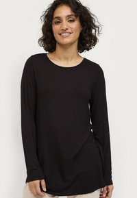 Vrouw met krullend haar in een zwarte shirt met lange mouwen, glimlachend, staande tegen een effen lichte achtergrond.