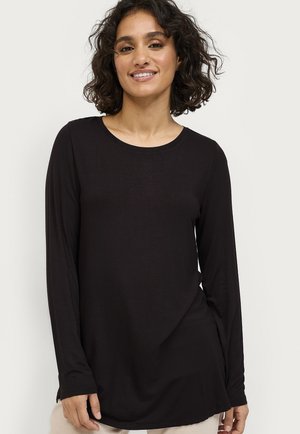 Vrouw met krullend haar in een zwarte shirt met lange mouwen, glimlachend, staande tegen een effen lichte achtergrond.