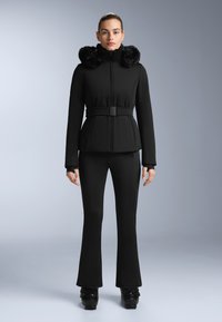 Černá lyžařská bunda s kapucí z umělé kožešiny, páskem v oblasti pasu a dlouhými rukávy v kombinaci s rozšiřujícími se černými lyžařskými kalhotami, obojí vyrobené z hladkého, voděodolného materiálu.