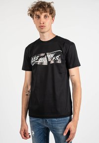 Camisa de algodón negra con un estampado gráfico que incluye el texto "LES HOMMES" en plata metálica, mangas cortas y un escote redondo.