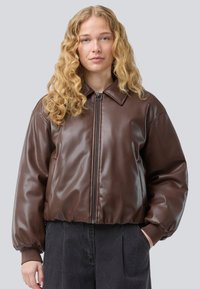 Hnedá umelá kožená bomber bunda so zipsom vpredu, širokým golierom, objemnými rukávmi a rebrovanými manžetami. Nosená cez tmavé nohavice.