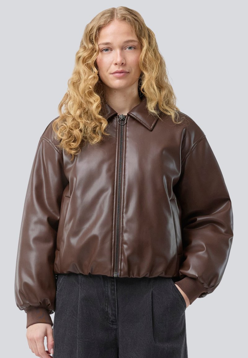 Hnedá umelá kožená bomber bunda so zipsom vpredu, širokým golierom, objemnými rukávmi a rebrovanými manžetami. Nosená cez tmavé nohavice.