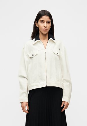 Chaqueta vaquera blanca con cuello negro, cierre frontal de cremallera, dos bolsillos en el pecho y diseño cropped, llevada sobre una falda negra plisada.