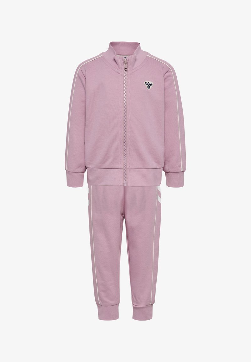 Jasnoróżowy dres z kurtką zapinaną na zamek i spodniami. Posiada białe paski jako akcenty i małe logo na piersi. Miękka faktura materiału.