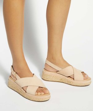 Pieds portant des sandales compensées beiges avec des brides croisées et une boucle à la cheville sur un fond clair.