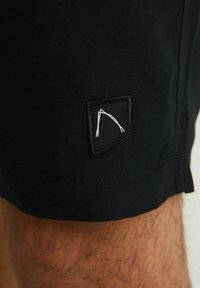Černé šortky s texturovanou tkaninou, které na boku zdobí malé černobílé vyšívané logo, vyzdvihující geometrický design.