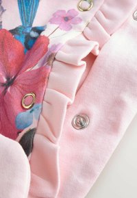 Rosa Stoff mit floralem Druck, verziert mit Rüschen am Rand und zwei Druckknöpfen: einem silbernen und einem goldenen.