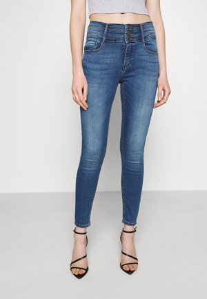 Slim-fit blå denim-jeans med tre-knaps lukning båret af en person, der står, kombineret med sorte rem-sandaler med høje hæle.