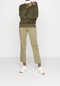 Maglione verde oliva con stampa animale e logo, abbinato a pantaloni chiari color khaki. Sneakers bianche con accenti beige completano l'outfit.