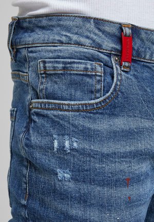 Primer plano de unos vaqueros de denim azul con parches desgastados y costuras naranjas, con una etiqueta roja en el lazo del cinturón y una camisa blanca metida dentro.