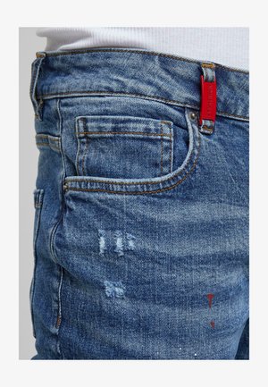 Ripresa ravvicinata di jeans in denim blu con toppe consumate e cuciture arancioni, dotati di un'etichetta rossa sul passante della cintura e una camicia bianca inserita.