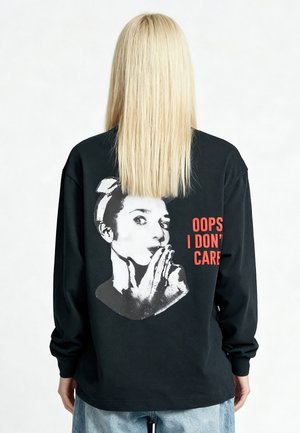Persona bionda che indossa una maglietta nera a maniche lunghe con una grafica di donna e la scritta "OOPS I DON'T CARE" sulla schiena, in piedi su sfondo bianco.