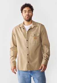 Camisa de manga larga beige de algodón, con un bolsillo frontal que lleva una etiqueta, cierre de botones y un corte relajado. Llevada sobre una camiseta blanca.